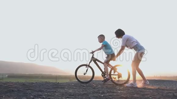 一位年轻的母亲教儿子在日落或日出的夏天骑自行车一个女人骑着自行车视频的预览图