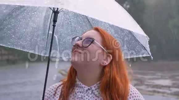 戴着眼镜的肥姜姑娘在雨中的公园里散步在天空中撑着伞看着视频的预览图