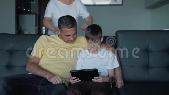 父亲母亲和儿子坐在沙发上看电影在平板电脑上玩游戏一个幸福的家庭视频的预览图
