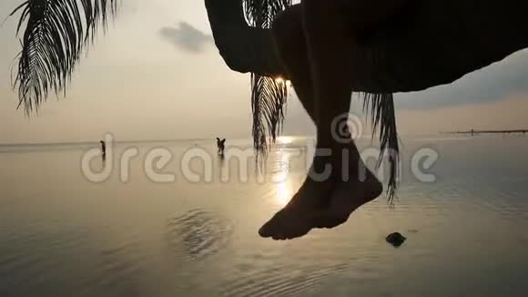 戴太阳镜的年轻人坐在棕榈树上在海岸日落时挥舞着双腿1920x1080视频的预览图