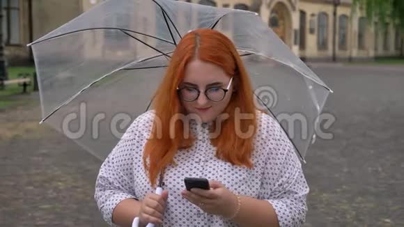 戴着眼镜的胖姜女孩正在公园里的智能手机上键入信息在雨天微笑着撑着伞视频的预览图