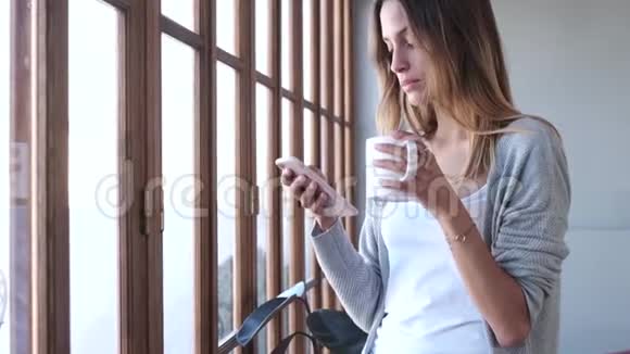漂亮的年轻女人在家客厅的窗户附近喝咖啡时用手机视频的预览图