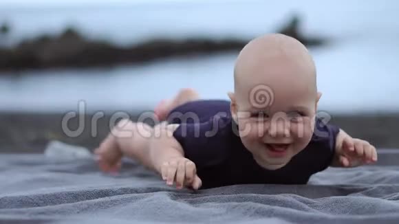 婴儿趴在海边的黑沙上笑着看着摄像机视频的预览图