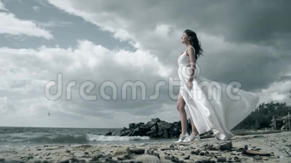 孤独美丽的女人穿着白色的裙子站在海洋上女孩手里拿着一件衣服在风中缓慢地飘动视频的预览图