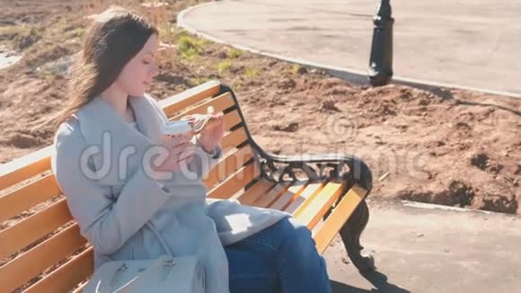年轻的黑发女人穿着外套坐在城市公园的长凳上吃着糖的面包喝着咖啡春天视频的预览图