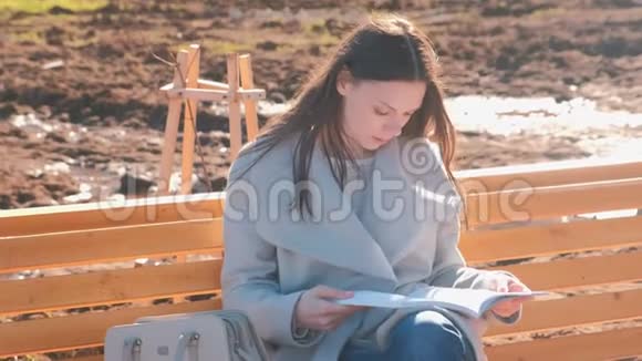 年轻漂亮的黑发女子学生坐在春天公园的长凳上读着一本书视频的预览图