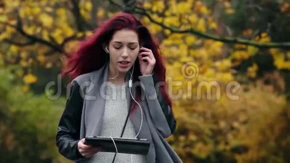 年轻的红发女商人在秋天的公园散步并通过平板电脑交流年轻漂亮的女人视频的预览图
