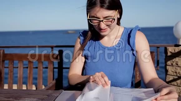 一个年轻的女孩坐在露台上的餐馆里俯瞰大海并检查菜单休闲海洋食物视频的预览图