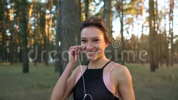 靠近微笑的女人把耳机插入耳朵在日落时穿过一片秋林视频的预览图