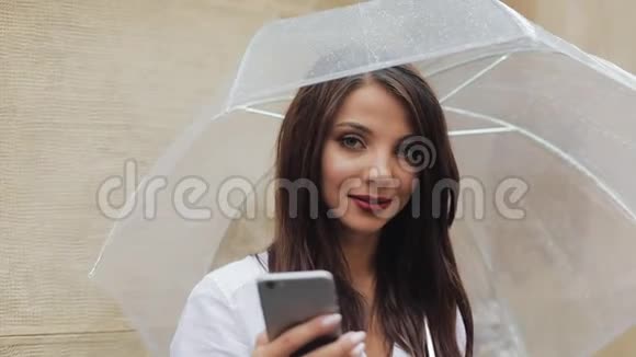 美丽的年轻商务女性在雨天的街道上使用智能手机微笑着拿着雨伞她看上去视频的预览图