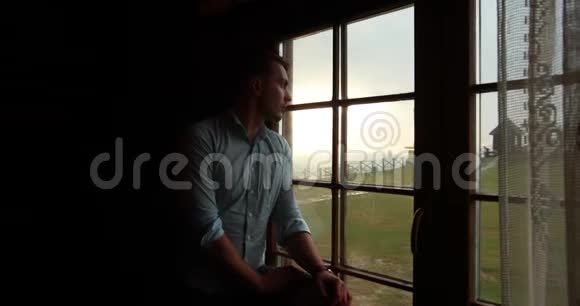 孤独在山里英俊的年轻人站在窗前看着史诗般的日落视频的预览图