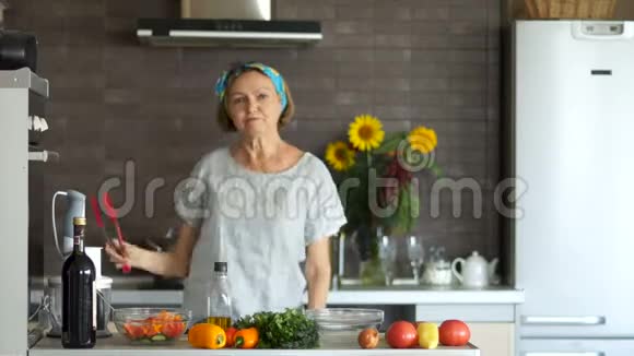 穿着整齐漂亮的中年妇女养老金领取者在厨房里跳舞用红色的铲子煎明亮的厨房视频的预览图