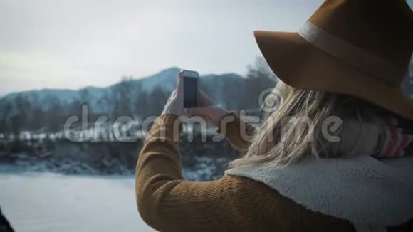 一个年轻的女孩旅行者在智能手机上拍照拍摄一条冰冻的冬季河流一顶帽子和一个旅行者背包视频的预览图