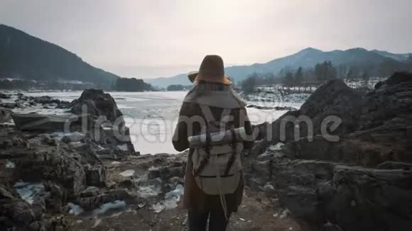 一个年轻的女孩旅行者在智能手机上拍照拍摄一条冰冻的冬季河流一顶帽子和一个旅行者背包视频的预览图