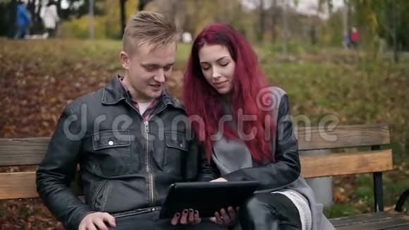 一位年轻女性红头发穿着皮夹克坐在公园的在公园的长凳上用一个数码塔贝视频的预览图