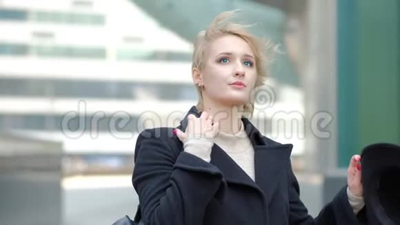 漂亮的金发女孩穿着黑色外套在模糊的现代建筑中摆出相机的姿势近距离观看慢镜头视频的预览图