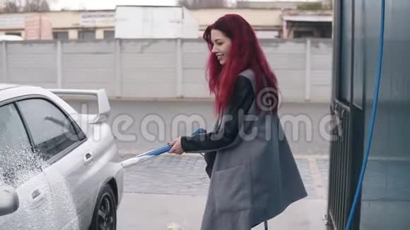 一位红头发的年轻女士在她的银色跑车上涂上泡沫并在自助洗车上涂上特殊的喷气式飞机视频的预览图