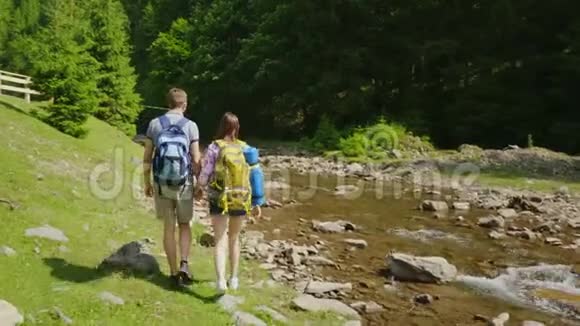一对年轻的游客在绿色森林的背景下沿着一条山河散步旅行和积极活动视频的预览图
