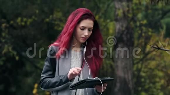 年轻漂亮的红发女人在秋天公园散步通过平板电脑听音乐年轻漂亮的女人视频的预览图