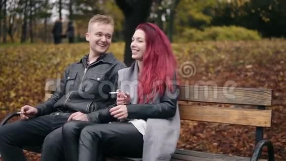 年轻迷人的一对夫妇坐在秋天公园的长凳上聊天有红发魅力的女人抽烟视频的预览图