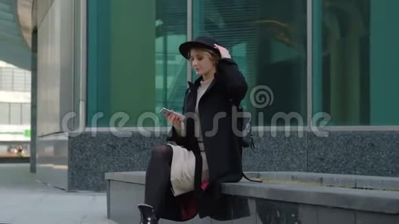 漂亮的金发女孩穿着黑色外套和帽子用智能手机耳机听音乐对抗城市街头慢动作视频的预览图