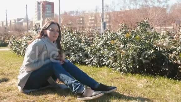年轻漂亮的黑发女人坐在春天公园的草地上听电话里的音乐唱一首歌视频的预览图