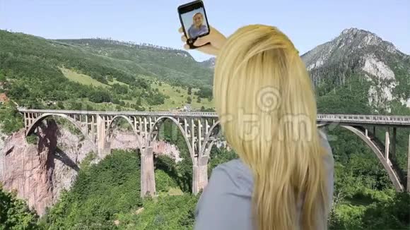 背景车拱桥上的金发美女在智能手机上自拍视频的预览图