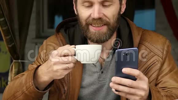 英俊的年轻人用智能手机坐在户外咖啡馆喝咖啡慢动作视频的预览图
