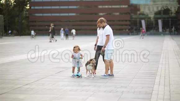 幸福家庭带着一只狗穿过城市广场穿裙子的小女孩骑着滑板车父亲带着一个视频的预览图