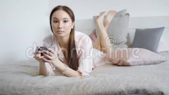 一位年轻漂亮的女士在床上喝了一杯热饮料她举着一个杯子穿着睡衣视频的预览图
