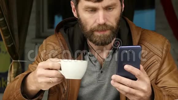 英俊的年轻人用智能手机坐在户外咖啡馆喝咖啡慢动作视频的预览图