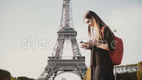 年轻漂亮的女人带着背包站在埃菲尔铁塔附近用智能手机浏览互联网视频的预览图