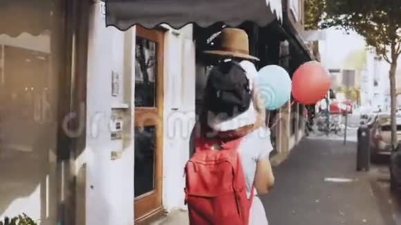 母亲背着儿子沿着街道走女孩带着一个戴着帽子和两个气球的小快乐的孩子走路4K视频的预览图