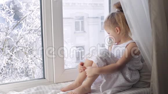 悲伤的小女孩透过窗户看着坐在窗台上外面冬天视频的预览图