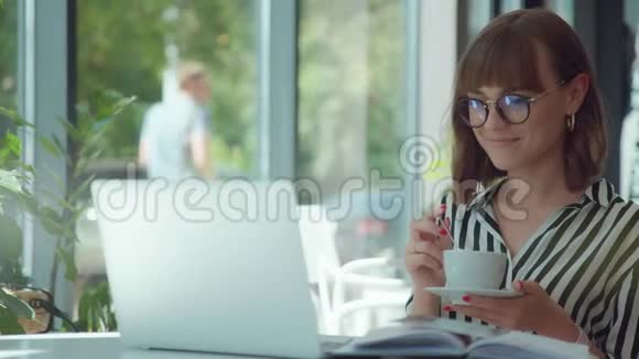 优雅的女人正在喝咖啡在笔记本电脑屏幕上看视频视频的预览图