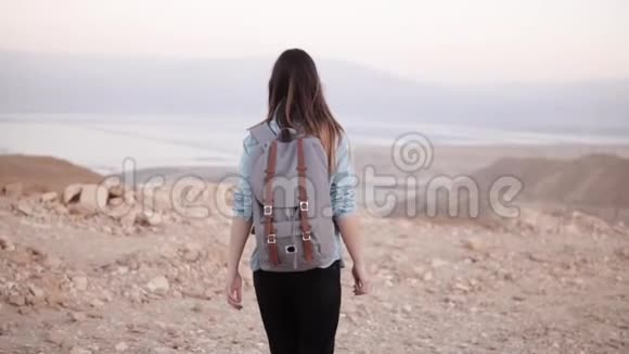 带背包的女人在沙漠里散步慢动作年轻女孩在干燥的沙子和岩石上游荡令人惊叹的山景视频的预览图