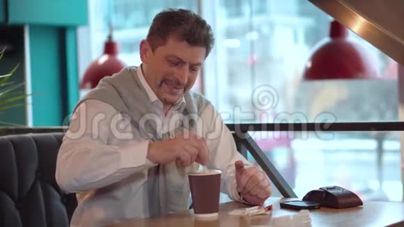 成熟的帅哥穿着一件白色衬衫穿着毛衣微笑着用棍子在刚做的早晨咖啡中搅动糖视频的预览图