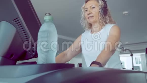 老年妇女在健身房的跑步机上订婚金发女性在跑步机上用耳机听音乐视频的预览图