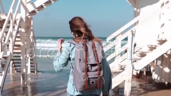 女孩走在白色楼梯到阳光明媚的海岸自由头发随风飘动带背包的女人在海滩上慢动作视频的预览图