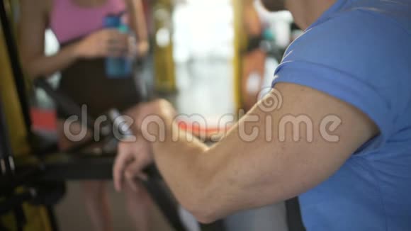 男健身房会员坐在设备上用运动水瓶与女士交谈视频的预览图