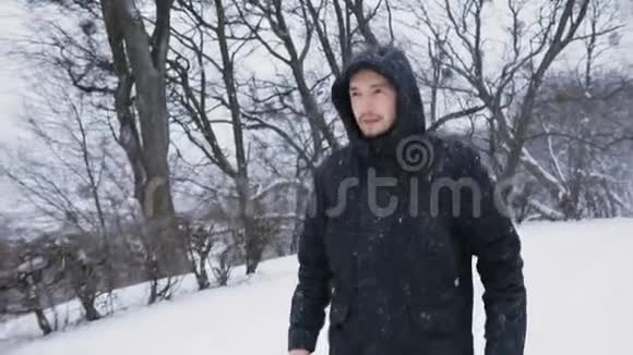 布鲁内特年轻男子戴着黑帽走在白雪皑皑的冬季公园里帽子擦着手寒冷的雪中中央视频的预览图