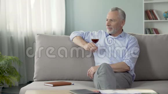 五十多岁的体贴的男人坐在沙发上享受红酒的味道视频的预览图