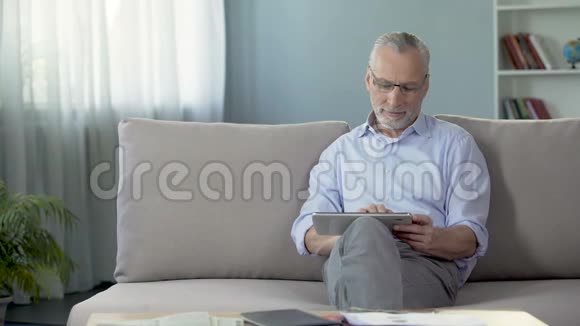 漂亮的高级男士坐在沙发上在平板电脑上滚动应用程序视频的预览图