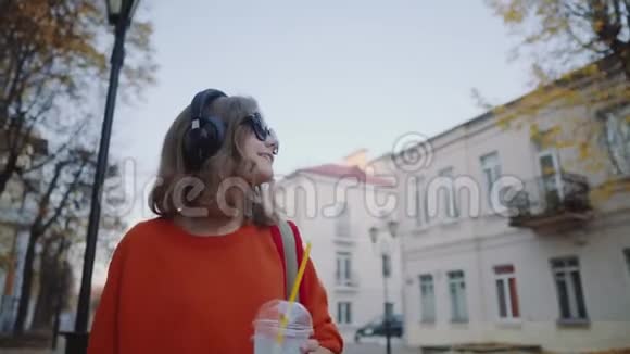 漂亮的时髦少年用耳机听音乐用塑料杯喝奶昔微笑着走在大街上视频的预览图