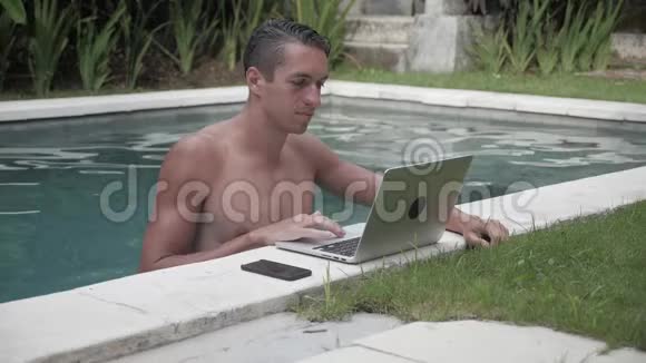 坐在游泳池里用笔记本电脑工作的年轻人他的自由职业者在游泳池的一个美丽的地方工作视频的预览图
