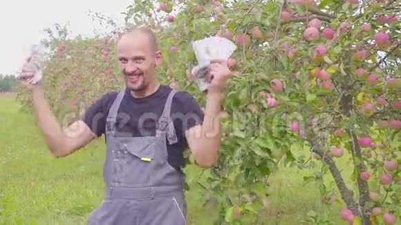 一个成功的快乐的年轻农民在苹果园和舞会上有很多钱商业成功的概念视频的预览图