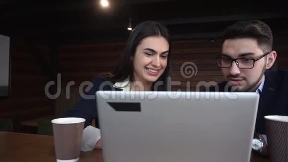 年轻的男人和开朗的女人正在看一台坐在咖啡馆里的笔记本电脑视频的预览图