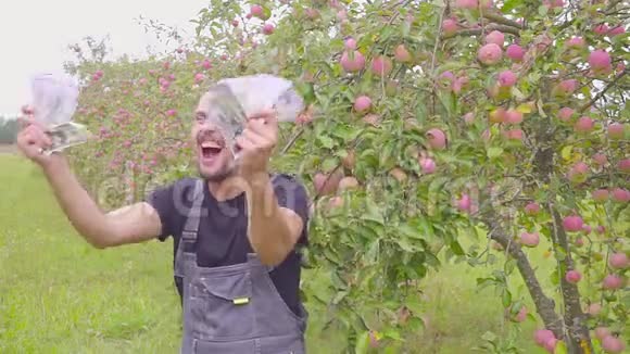 一个成功的快乐的年轻农民在苹果园和舞会上有很多钱商业成功的概念视频的预览图