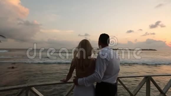度假时新娘和新郎在栏杆附近的热带海滩欣赏日落新婚夫妇拥抱视频的预览图