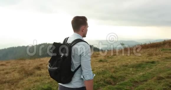 孤独在山里英俊的年轻人背着背包在灰色的天空下沿着山路散步视频的预览图